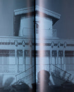 An open book or photo album showing a monochrome architectural photograph spanning two pages, with the text '1899-1935' in the top right of the right page. A hand is holding the left edge of the book against a light gray background.