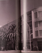 An open vintage art book held by a hand, showing a sepia-toned photograph of a 1935-1941 street scene with palm trees and a corner building. The book has gold-toned edges and is photographed against a light gray background.
